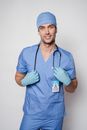 Positive man doctor in blue uniform and protective gloves standing with stethoscope and looking at camera against light background