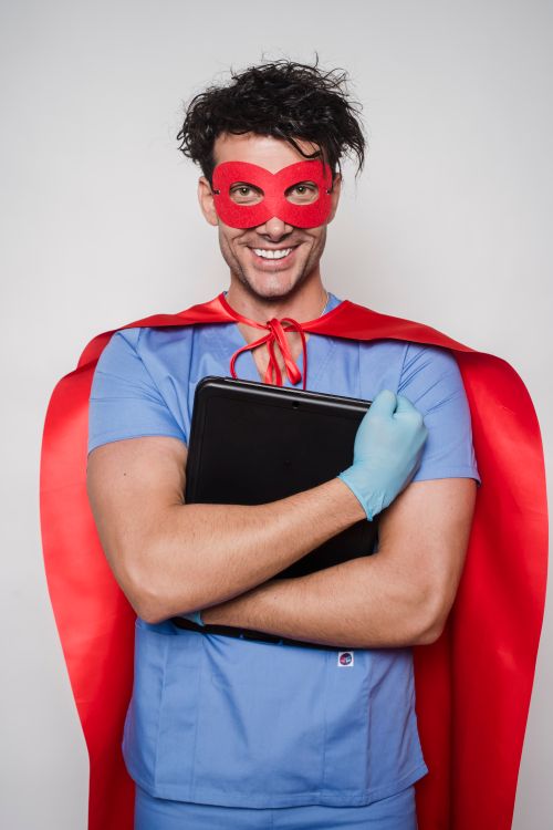 Free Cheerful man doctor in superhero costume with mask and cape standing with folder with documents and looking at camera against light wall Stock Photo