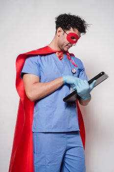 Pensive man doctor in medical uniform and superhero costume with mask and cape standing with clipboard against light background
