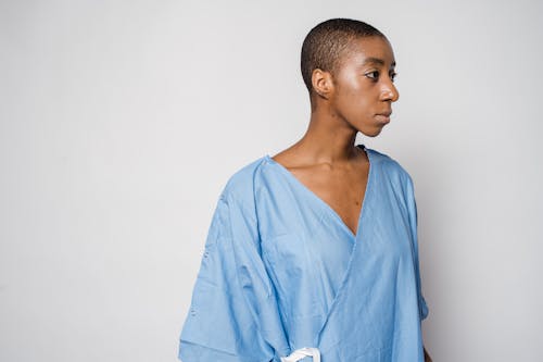 Pensive androgynous African American female patient with short dark hair in medical robe standing against light wall and looking away