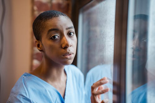 Thoughtful African American female with short dark hair in blue medical robe standing near window in room and looking at camera in daytime