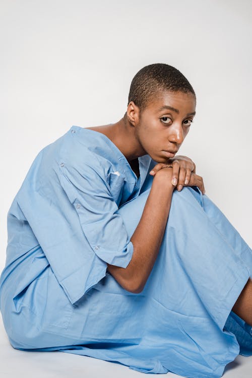 African American female patient in blue gown looking at camera while sitting on floor on white background