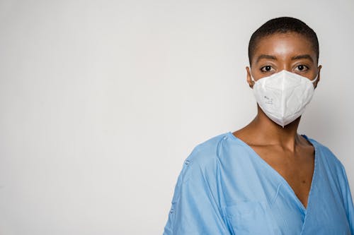 Serious African American female doctor in medical mask and blue uniform standing against white background