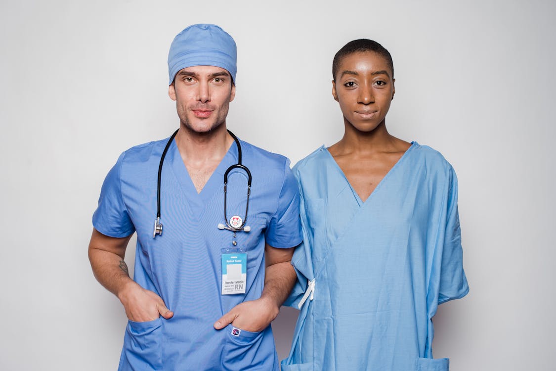 Free Positive male doctor in blue uniform with hands in pockets smiling and looking at camera while standing near nurse holding hands behind back in hospital Stock Photo