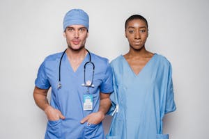 Positive male doctor in blue uniform with hands in pockets smiling and looking at camera while standing near nurse holding hands behind back in hospital
