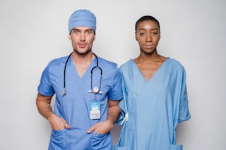 Positive male doctor in blue uniform with hands in pockets smiling and looking at camera while standing near nurse holding hands behind back in hospital