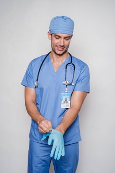 Positive man in professional medical uniform and stethoscope smiling while wearing latex gloves on gray wall background