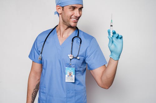 Smiling male doctor in uniform and gloves with stethoscope holding syringe while standing against white background in clinic