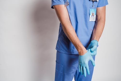 Crop anonymous male doctor in uniform with stethoscope putting on sterile gloves while standing near white wall in hospital