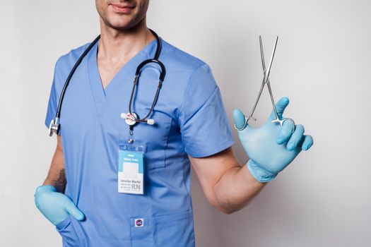 Crop faceless male doctor in blue medical uniform and gloves with stethoscope and medical forceps with id card on white background in bright studio