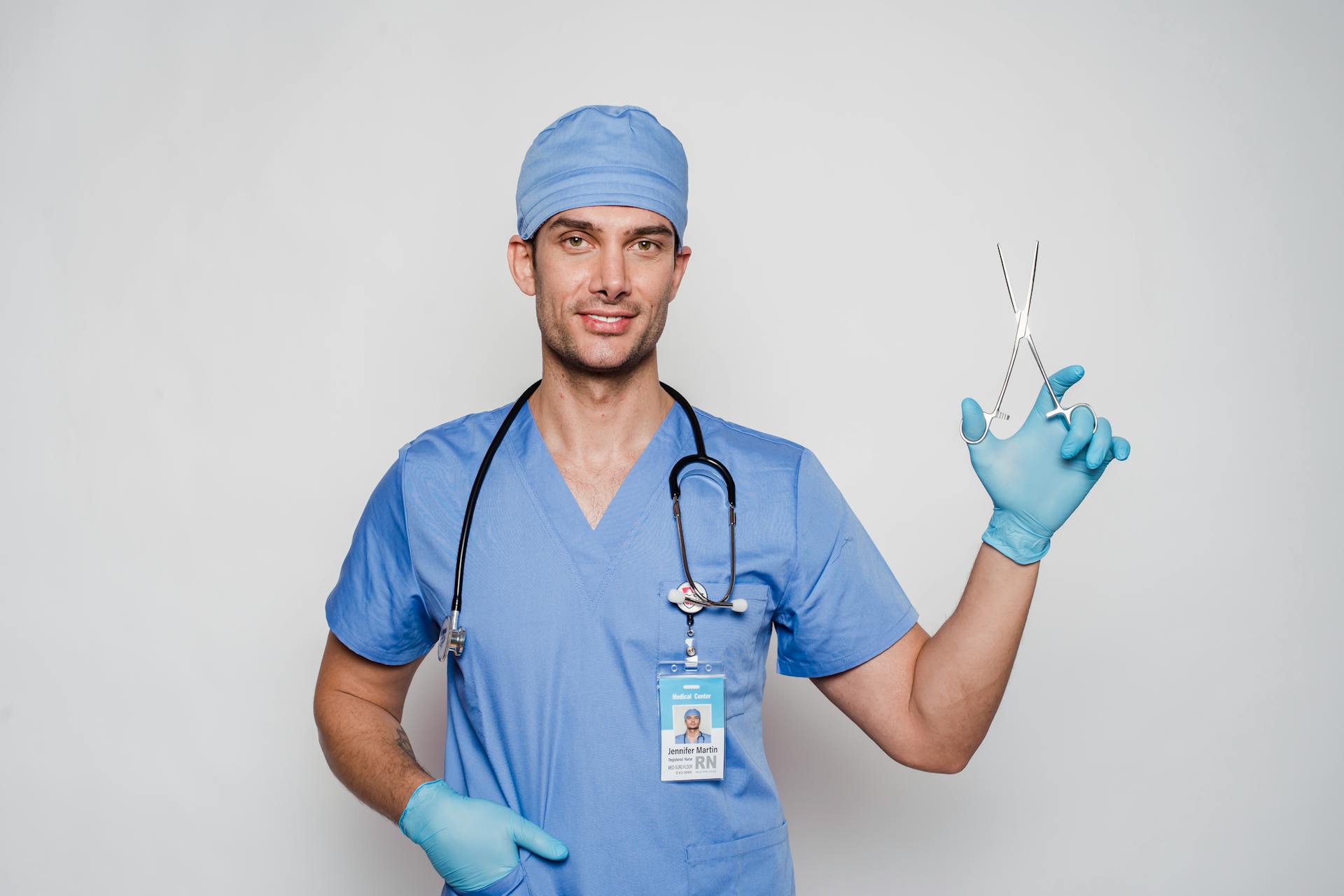 Positive adult doctor in blue uniform and cap with gloves and stethoscope with medical instrument and id card looking at camera on white background in light studio