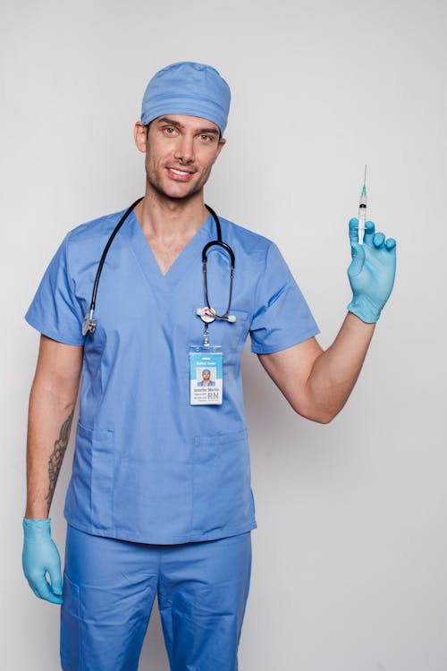 Smiling adult male doctor in blue medical uniform and gloves in cap with stethoscope and medical syringe with id card on white background looking at camera in bright studio