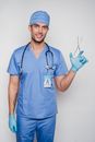 Positive adult doctor in blue uniform and cap with gloves and stethoscope with medical instrument and id card looking at camera in light studio on white background