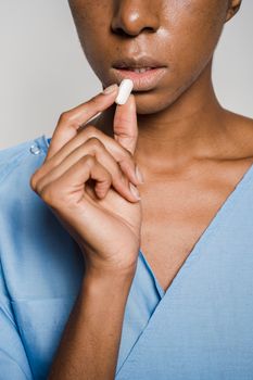 Crop faceless African American female patient wearing blue medical robe while taking white tablet in light studio on white background