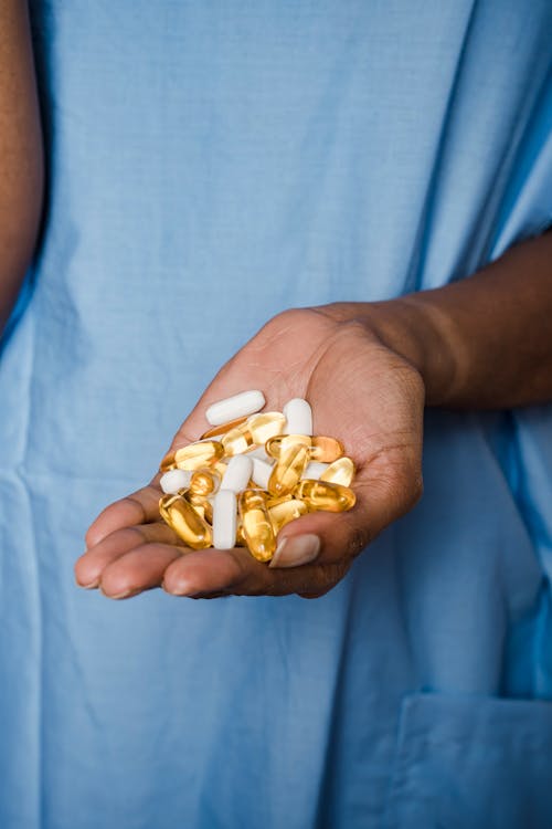 Crop unrecognizable black patient in blue medical robe demonstrating white and yellow medical pills in hand in bright room