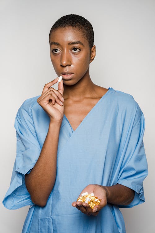 Pensive African American female patient with short hair in medical clothes eating painkillers and looking away against light wall in studio