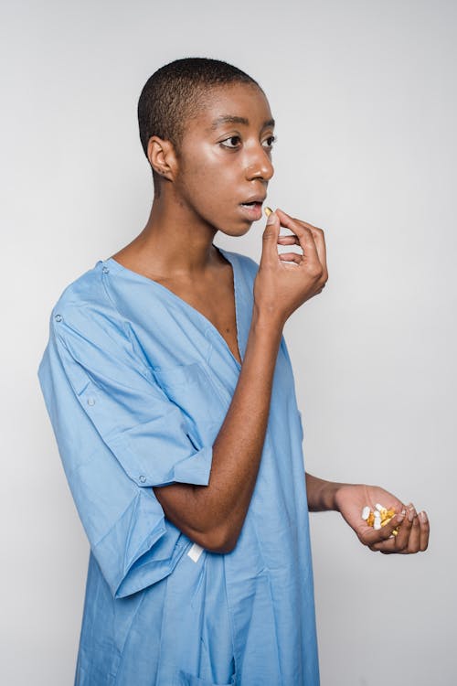 Pensive African American woman with short dark hair in blue medical robe standing with handful of medicines in hand and eating pill in room with light wall