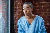 Pensive African American female patient with short dark hair in blue medical robe looking at window against brick wall in daylight