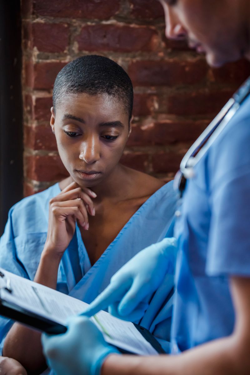 A Black woman in a hospital gown reviews a diagnosis on a clipboard while talking with her doctor in a hospital corridor, a moment that often begins with an abnormal blood count.