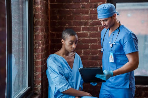 Thoughtful doctor in uniform and gloves showing diagnosis to African American female patient with short hair in blue medical robe in hallway of hospital in daytime