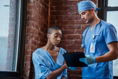 Calm doctor in blue uniform standing with clipboard and demonstrating diagnosis to African American woman in patient robe in hallway of clinic