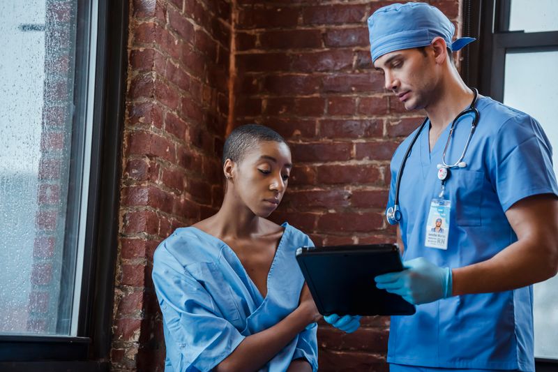 A Black woman patient in a blue medical gown stands beside a white male clinician in blue scrubs holding a tablet; both look at the tablet screen in an engaged conversation, hospital brick-wall context.