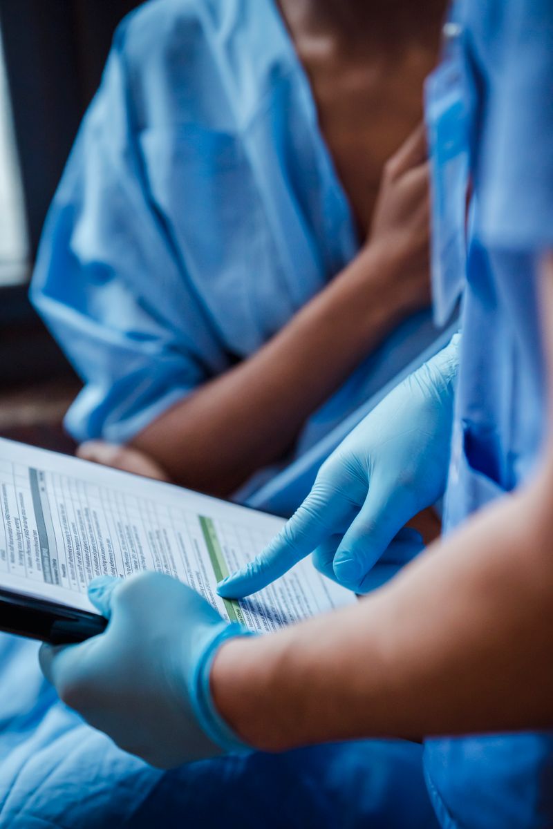 A clinician in scrubs and gloves shows a diagnosis on a clipboard to a Black woman patient during a clinic consultation after breast imaging.