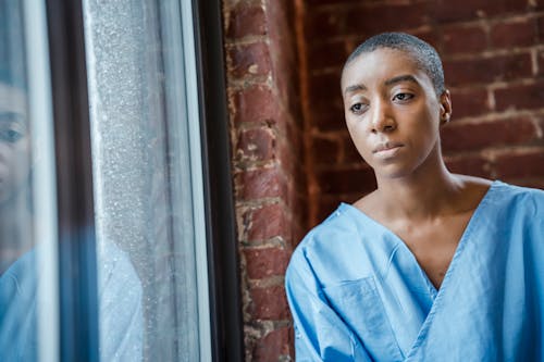 Pensive African American female patient with short dark hair in blue medical robe thoughtfully looking at window against brick wall in daytime