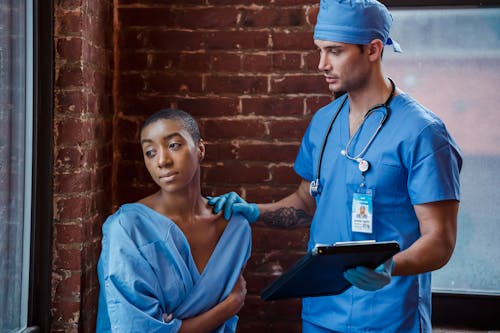 Man doctor in blue uniform and sterile gloves with stethoscope demonstrating diagnosis to thoughtful African American female patient in corridor of clinic in daytime