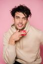 Charismatic man with curly dark hair in stylish clothes sitting with red condom in hand and looking at camera in studio against pink background