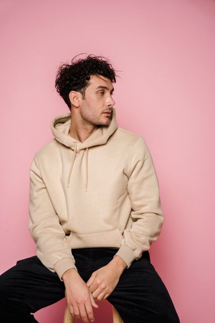 Thoughtful male with dark hair in stylish clothes sitting on wooden chair and looking away in studio against pink background