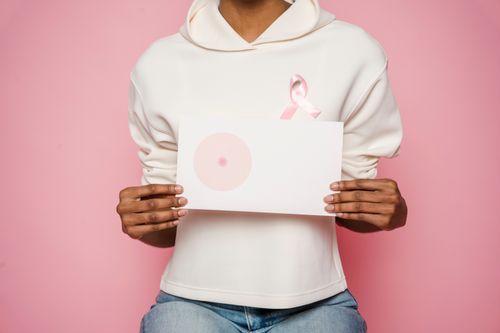 Free Crop anonymous African American woman in light hoodie with pink ribbon showing paper with painted one breast during Breast Cancer Awareness Month in studio with pink background Stock Photo