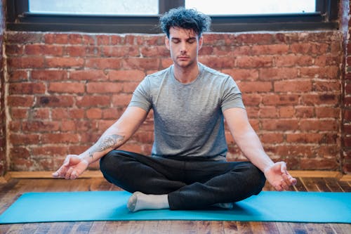 穿著灰色圓領t恤和黑色的褲子，坐在藍色瑜伽墊上的男人