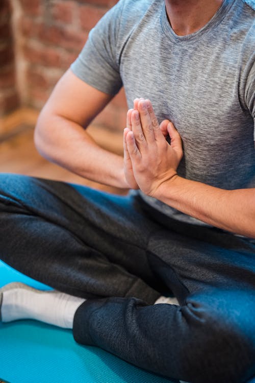 High angle of crop faceless male sitting with crossed legs in Sukhasana with praying hands on chest