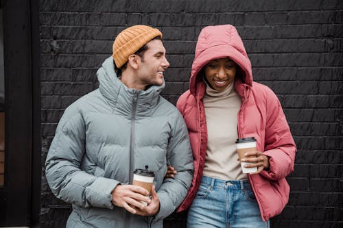 Free Positive multiethnic couple in outerwear standing close with cups of takeaway coffee on street Stock Photo