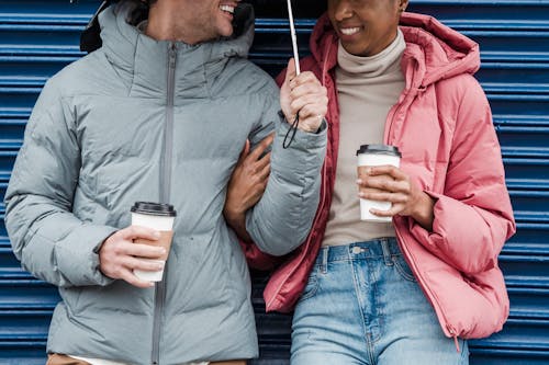 Free Crop smiling multiethnic couple wearing warm outerwear standing together with coffee cups in rainy day Stock Photo