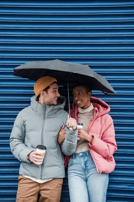 Happy multiethnic couple in outerwear standing with cups of takeaway coffee under umbrella and looking at each other with love