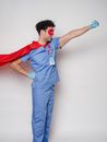 Positive male doctor in blue uniform with name tag under flying cape standing with hand on waist and fist forward against gray background