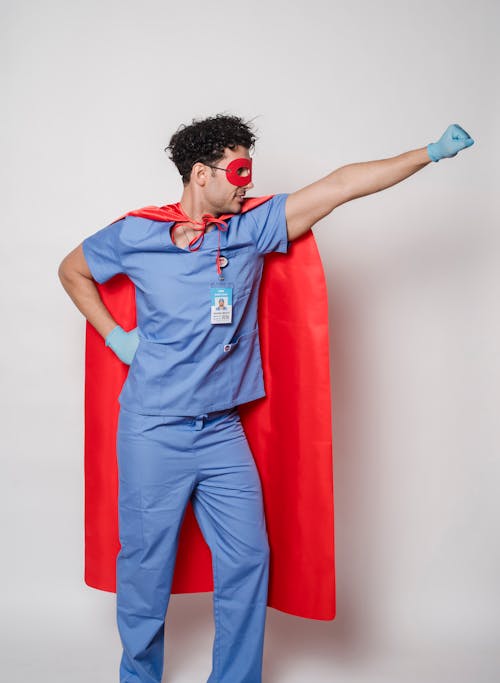 Male medical practitioner wearing superhero cape and uniform standing with hand on waist and raised fist for fighting against coronavirus
