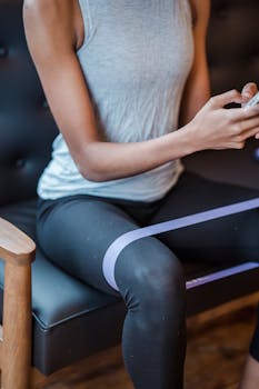 A woman in activewear uses a smartphone on a couch with a resistance band for exercises.