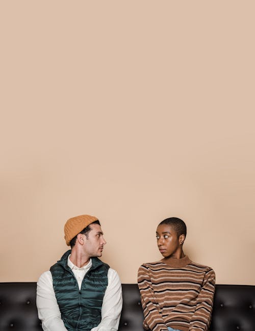 Misunderstanding multiethnic couple having problems in relationship sitting on leather couch and looking at each other against beige background