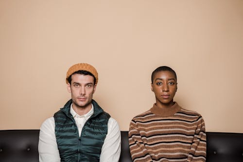 Emotionless multiethnic couple sitting on couch against beige background and looking at camera