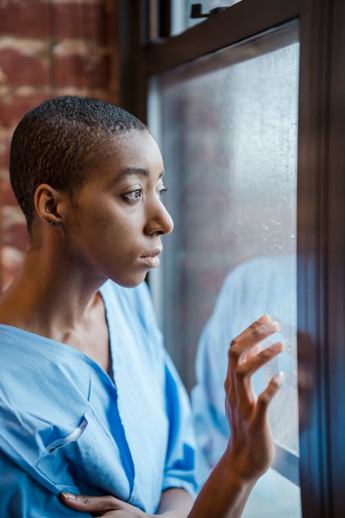 Free Crop unhappy sick African American bald female touching glass of wet window in hospital Stock Photo