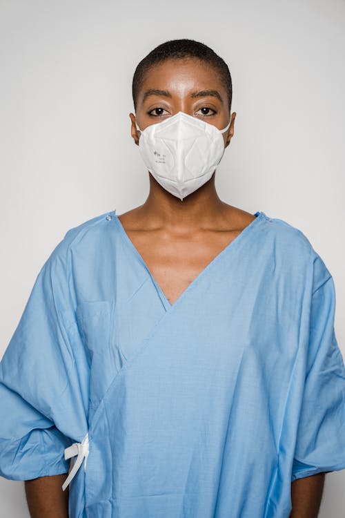 Bald African American anonymous female in blue patient gown and medical mask looking at camera on white background