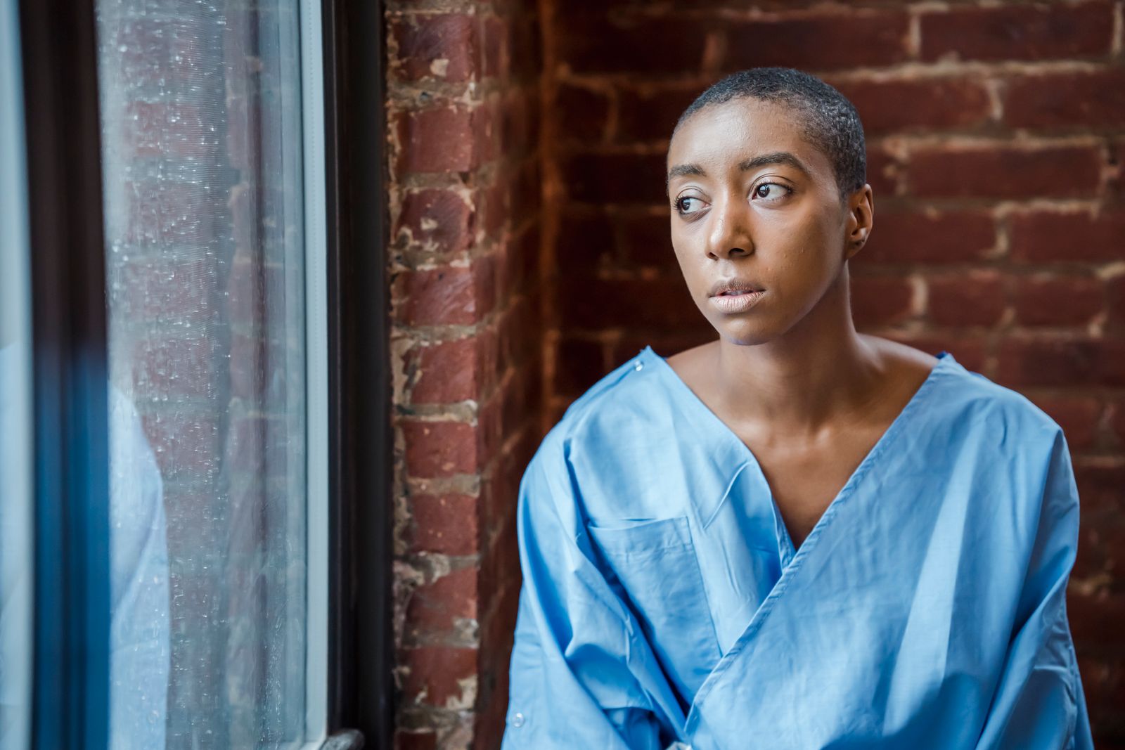 A young Black woman in a blue hospital patient gown stands against a brick wall in a clinical setting, looking out a window.