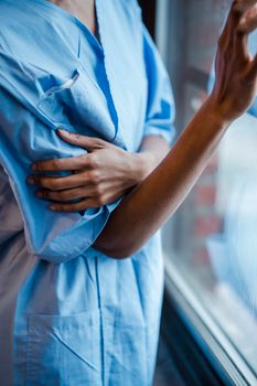 Crop anonymous ill African American female in blue patient gown near window of hospital