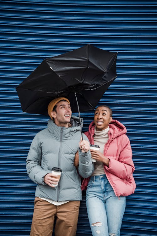 Diverse excited man and woman in outer weather under umbrella caught in gust of wind near blue wall on street