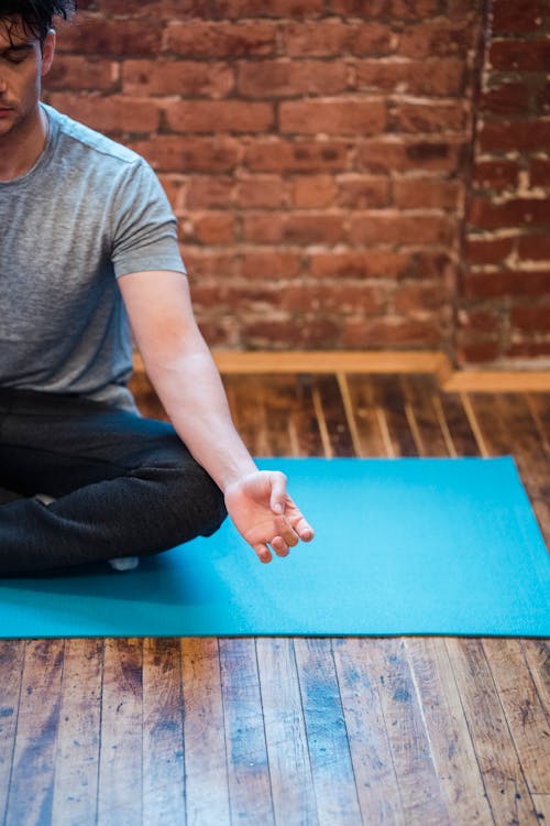 Crop concentrated male doing yoga and meditating on blue mat on timber floor at home