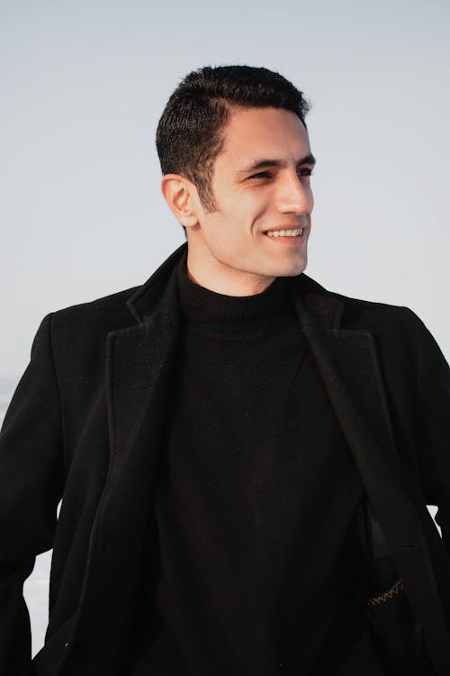 Cheerful young male in stylish black coat and warm turtleneck with hands in pockets looking away