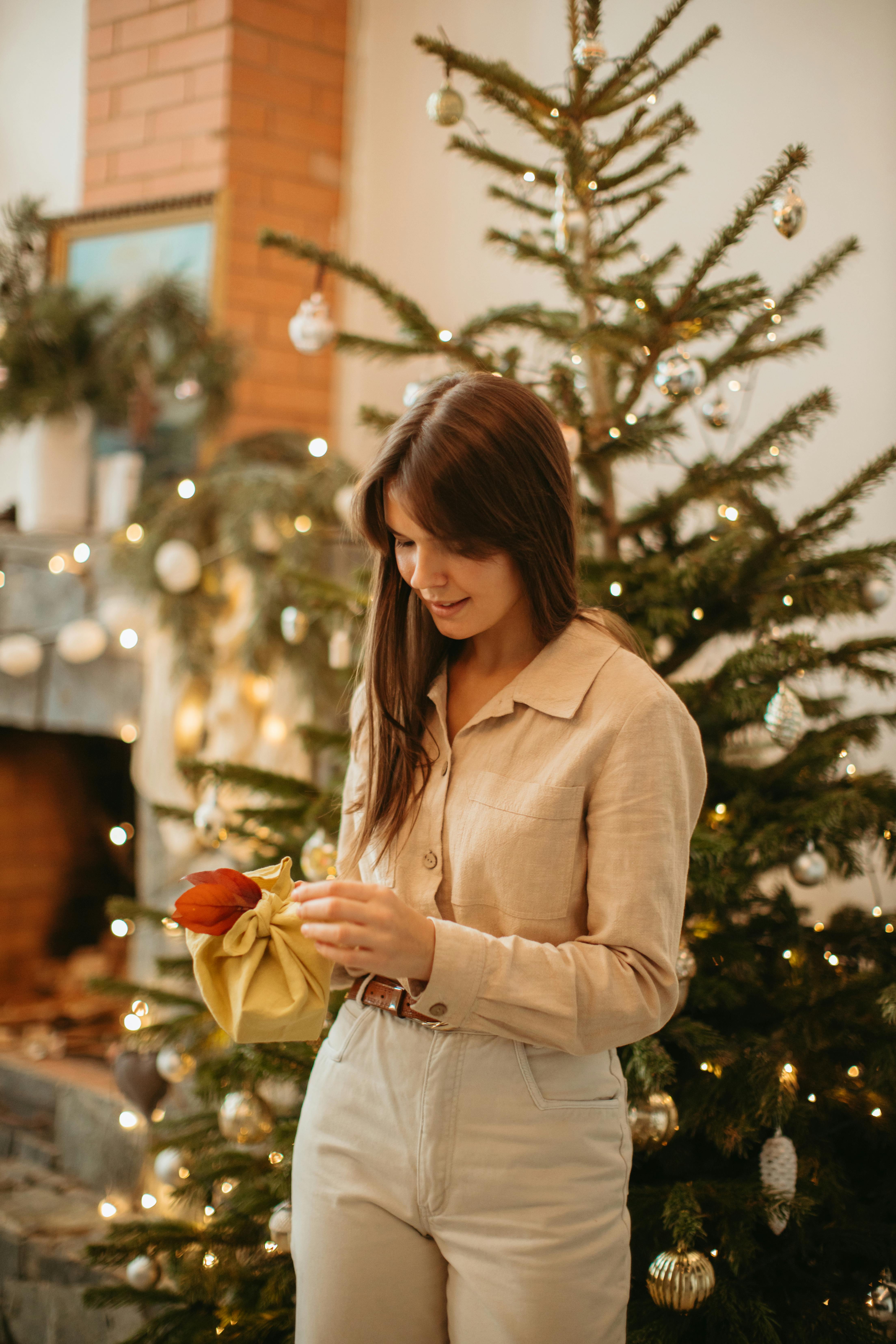 A woman standing by a Christmas tree, holding a present, surrounded by holiday decorations indoors.